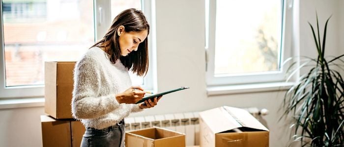 woman in white sweater and jeans looking at clipboard surrounded by brown moving boxes in room with windows and green plant